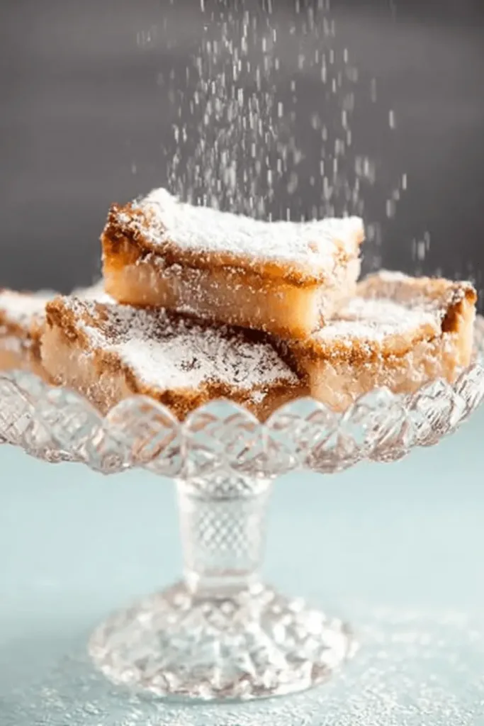 Golden gooey butter cake bars on a decorative glass stand with powdered sugar being sifted over them.